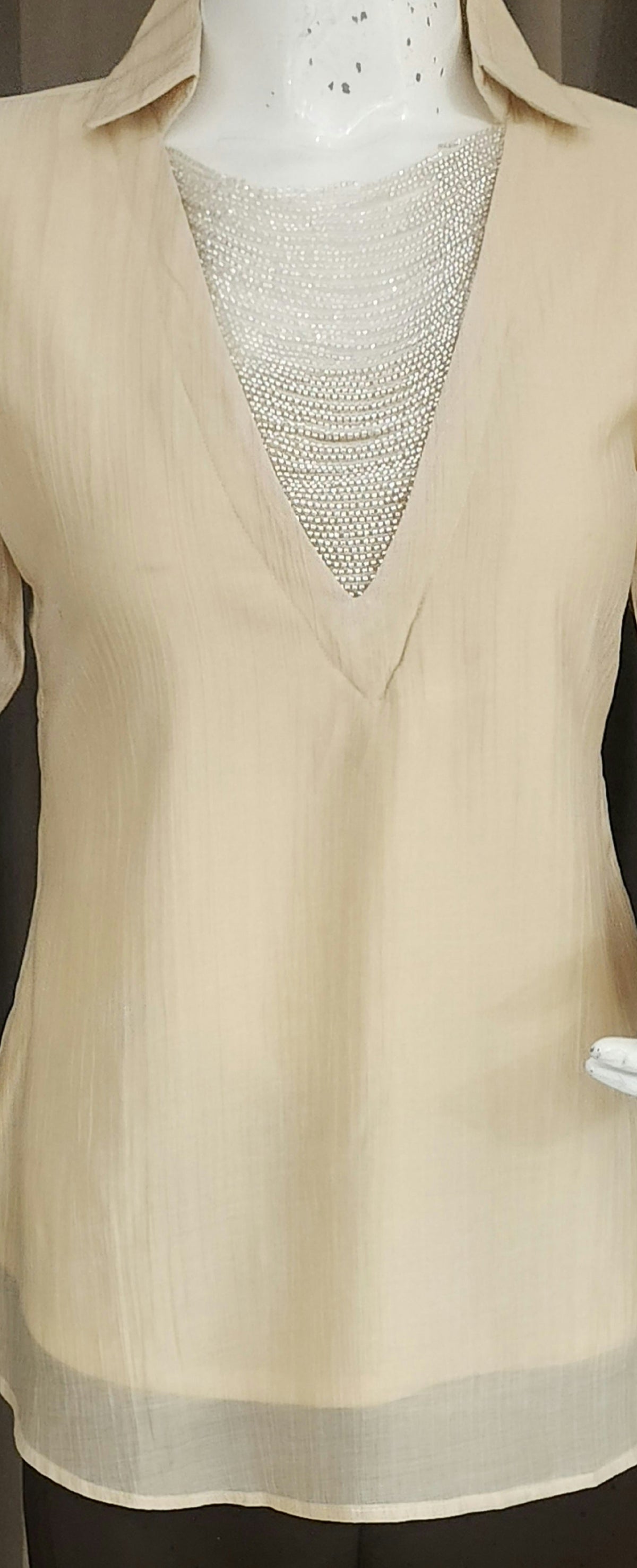 A beige long-sleeve blouse with a V-neck and sheer underlayer, displayed on a mannequin with dark brown wide-leg pants against a brown curtain backdrop.
